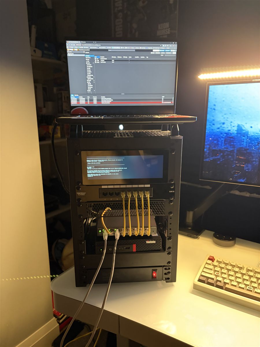 Front of a small server rack with a patch panel, managed switch and mini PC, and a laptop on top showing Proxmox